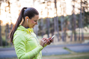 A young female runner with earphones outdoors in autumn nature, using smartphone. Copy space. - HPIF27305