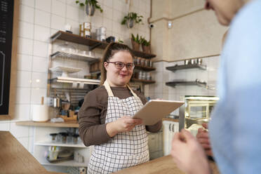 Smiling cafe owner with down syndrome taking order of customer through tablet PC in coffee shop - ABIF02001