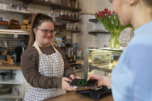 Lächelnder Café-Besitzer mit Down-Syndrom und Kunde beim Bezahlen mit dem Smartphone - ABIF02011