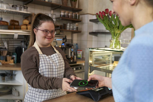 Smiling cafe owner with down syndrome and customer making payment through smart phone - ABIF02011