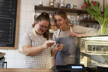 Smiling cafe owner with down syndrome learning cash register in coffee shop - ABIF02024