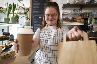 Glücklicher Café-Besitzer mit Down-Syndrom hält Papiertüte und Einweg-Kaffeebecher im Café - ABIF02030