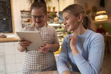 Lächelnder Café-Besitzer mit Down-Syndrom nimmt die Bestellung eines Kunden über einen Tablet-PC im Café auf - ABIF02034