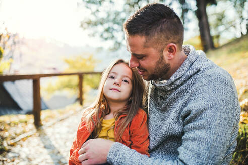 Ein junger Vater hält seine kleine Tochter in der herbstlichen Natur, sitzend. - HPIF27474