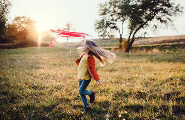 Ein glückliches kleines Mädchen spielt mit einem Regenbogen-Handdrachen in der herbstlichen Natur bei Sonnenuntergang. - HPIF27499