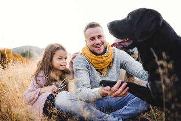 Vater mit kleiner Tochter und schwarzem Hund im Gras sitzend in herbstlicher Natur. - HPIF27518