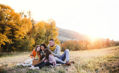 Ein Porträt von glücklichen jungen Familie mit zwei kleinen Kindern sitzen auf einem Boden im Herbst Natur bei Sonnenuntergang, mit Picknick. - HPIF27524
