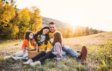 Ein Porträt von glücklichen jungen Familie mit zwei kleinen Kindern sitzen auf einem Boden im Herbst Natur bei Sonnenuntergang, mit Picknick. - HPIF27525