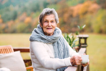 Eine ältere Frau mit einer Tasse Kaffee steht an einem sonnigen Herbsttag auf einer Terrasse im Freien. - HPIF27615