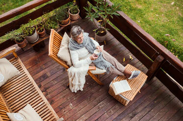 A top view of elderly senior woman with a cup of coffee and a book sitting outdoors on a terrace on a sunny day in autumn, feet on a basket. - HPIF27623