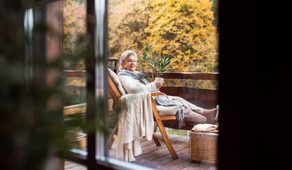 Eine ältere Frau sitzt an einem sonnigen Herbsttag auf einer Terrasse im Freien, aufgenommen durch Glas. - HPIF27628