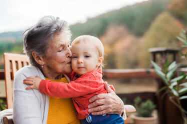 An elderly woman kissing a toddler great-grandchild on a terrace on a sunny day in autumn. - HPIF27643