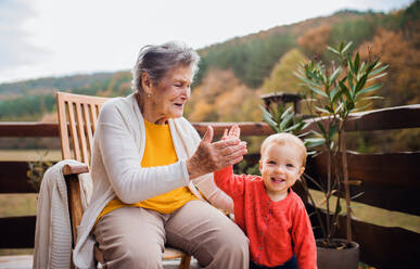 An elderly woman with a toddler great-grandchild on a terrace on a sunny day in autumn, giving high five. - HPIF27647