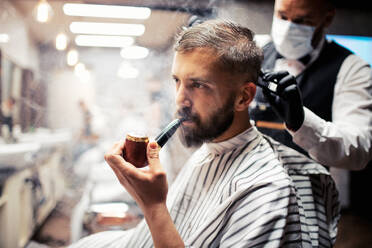 Gut aussehender Hipster-Kunde, der einen Friseur und einen Haarstylisten in einem Friseursalon besucht und dabei eine Pfeife raucht. - HPIF27659