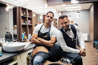 Two male haidressers and hairstylists sitting in barber shop, posing for a photograph. - HPIF27693