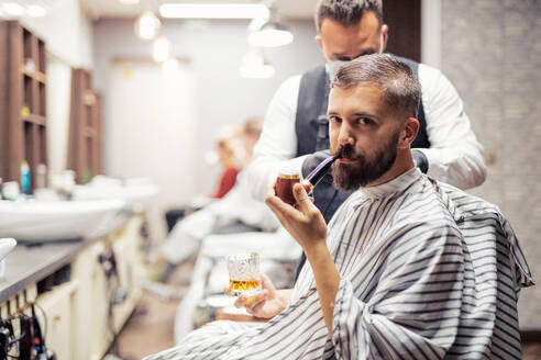 Gut aussehender Hipster-Kunde, der einen Friseur und einen Haarstylisten in einem Friseursalon besucht und dabei eine Pfeife raucht. - HPIF27705