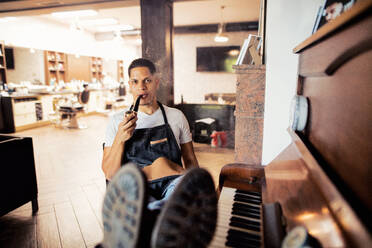 Young male hispanic haidresser and hairstylist sitting in barber shop, smoking a pipe and holding a drink. - HPIF27730