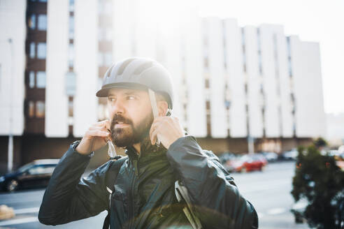 Männlicher Hipster-Kurier, der beim Ausliefern von Paketen in der Stadt einen Helm aufsetzt. - HPIF27741