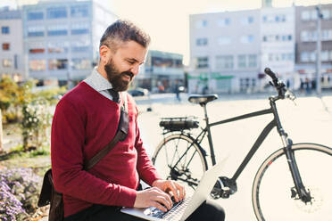 Hipster businessman commuter with bicycle on the way to work in city, sitting on a bench and using laptop. - HPIF27787