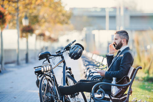 Seitenansicht eines Geschäftsmannes mit Smartphone und Fahrrad, der auf einer Bank in der Stadt sitzt und ein Telefonat führt. - HPIF27805