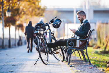 A businessman commuter with bicycle sitting on bench in city and using smartphone when traveling home from work. - HPIF27806