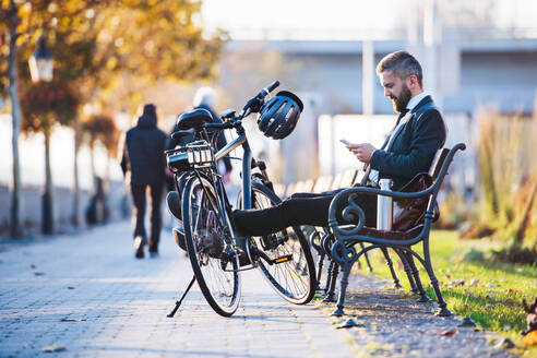 Ein Geschäftsmann mit Fahrrad sitzt auf einer Bank in der Stadt und benutzt sein Smartphone auf dem Heimweg von der Arbeit. - HPIF27806