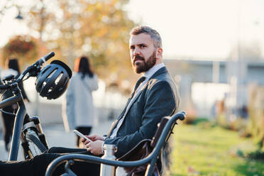 Seitenansicht eines Geschäftsmannes, der mit Smartphone und Fahrrad auf einer Bank in der Stadt sitzt. - HPIF27807