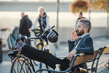 A side view of businessman commuter with smartphone and bicycle sitting on bench in city, making a phone call. - HPIF27808