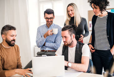 Eine Gruppe junger, fröhlicher Geschäftsleute mit Laptop sitzt und steht um einen Tisch in einem modernen Büro und hält eine Besprechung ab. - HPIF27874