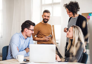 Eine Gruppe junger, fröhlicher Geschäftsleute mit Laptop sitzt und steht um einen Tisch in einem modernen Büro und hält eine Besprechung ab. - HPIF27878