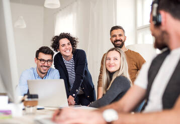 Eine Gruppe junger, fröhlicher Geschäftsleute mit Laptop sitzt und steht um einen Tisch in einem modernen Büro und hält eine Besprechung ab. - HPIF27880