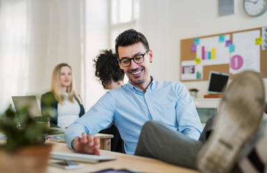 Ein Porträt eines jungen Geschäftsmannes mit Kollegen in einem modernen Büro, Füße auf dem Tisch. - HPIF27884