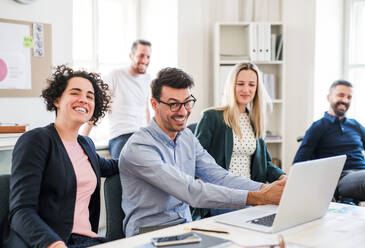 Eine Gruppe junger, fröhlicher Geschäftsleute mit Laptop sitzt und steht um einen Tisch in einem modernen Büro und hält eine Besprechung ab. - HPIF27904