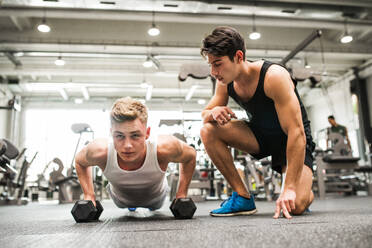 Young fit men friends in gym doing push ups on dumbbells, a friend supporting him. - HPIF27973