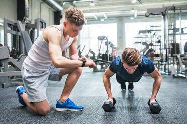 Young fit men friends in gym doing push ups on dumbbells, measuring time on smartwatch. - HPIF27975