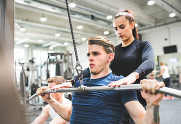 Junger, gut aussehender, fitter Mann, der an einer Pull-Down-Maschine im Fitnessstudio trainiert, ein Personal Trainer steht hinter ihm und hilft ihm. Bodybuilder, der mit einer Kabelgewichtsmaschine trainiert. - HPIF27978