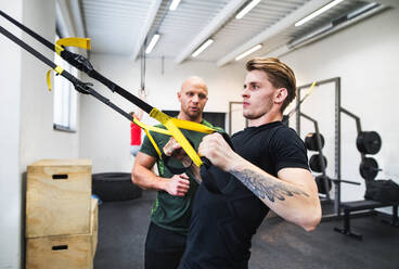 Ein fitter junger Mann mit einem Personal Trainer im Fitnessstudio, der mit TRX trainiert. - HPIF27986