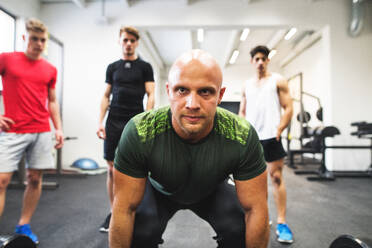 Drei fitte junge Männer betrachten einen Personal Trainer im Fitnessstudio, der eine Langhantel hebt. - HPIF27991