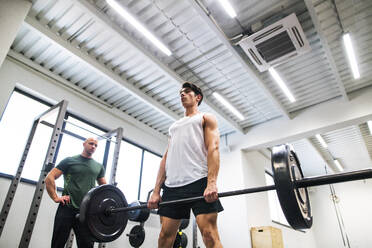 Young handsome fit man with a personal trainer in gym working out, lifting bar of barbell, flexing muscles. - HPIF27993