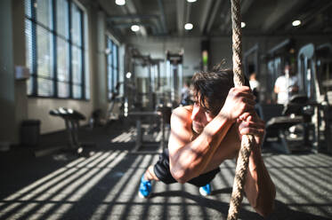 Junger, fitter Mann trainiert im Fitnessstudio, ein Junge hält sich im Schneidersitz an einem Kletterseil fest. - HPIF28023