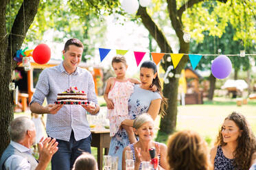 Familienfeier draußen im Garten. Große Gartenparty. Geburtstagsfeier. Ein junger Mann hält eine Geburtstagstorte. - HPIF28087