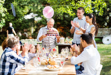 Familienfeier draußen im Garten. Große Gartenparty. Geburtstagsfeier. Ein älterer Mann hält eine Geburtstagstorte. - HPIF28088