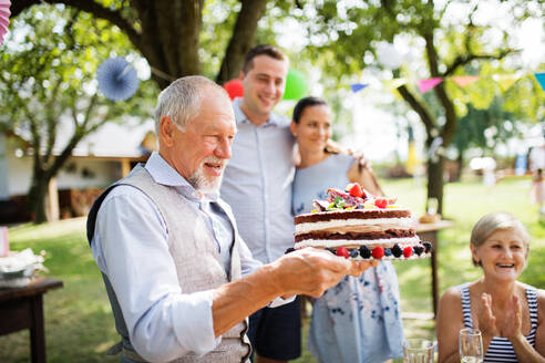 Familienfeier draußen im Garten. Große Gartenparty. Geburtstagsfeier. Ein älterer Mann hält eine Geburtstagstorte. - HPIF28089