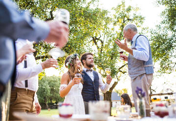Ein älterer Mann hält eine Rede bei einem Hochzeitsempfang draußen im Hintergrund. Braut und Bräutigam und andere Gäste halten Gläser. - HPIF28124