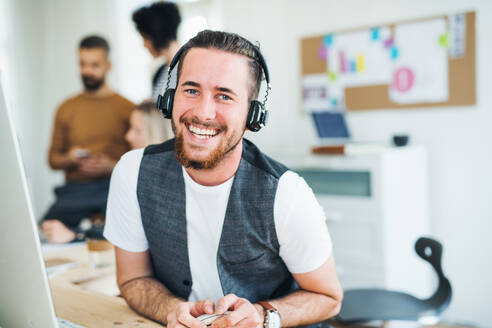 Ein Porträt eines jungen Geschäftsmannes mit Kopfhörern und Kollegen in einem modernen Büro, lachend. - HPIF28150