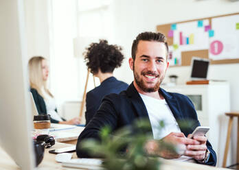 A portrait of young hipster businessman with smartphone and colleagues in the background in a modern office. - HPIF28166