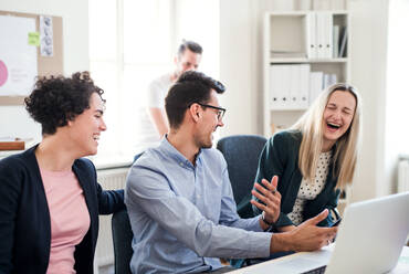 Eine Gruppe junger, fröhlicher Geschäftsleute mit Laptop sitzt in einem modernen Büro am Tisch und lacht bei einer Besprechung. - HPIF28179