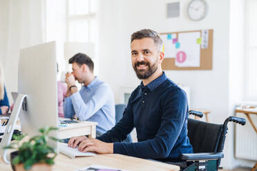 Junger Geschäftsmann im Rollstuhl mit Kollegen, die in einem modernen Büro arbeiten und einen Computer benutzen. - HPIF28180