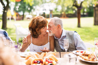 Ein älteres Paar sitzt bei einer Gartenparty am Tisch und berührt die Nasen. Familienfeier im Garten. - HPIF28199