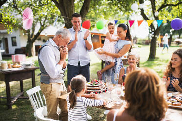 Ein älterer Mann mit seiner Großfamilie, der vor Glück weinend auf die Geburtstagstorte blickt. Feier im Freien im Hinterhof oder große Gartenparty. - HPIF28200